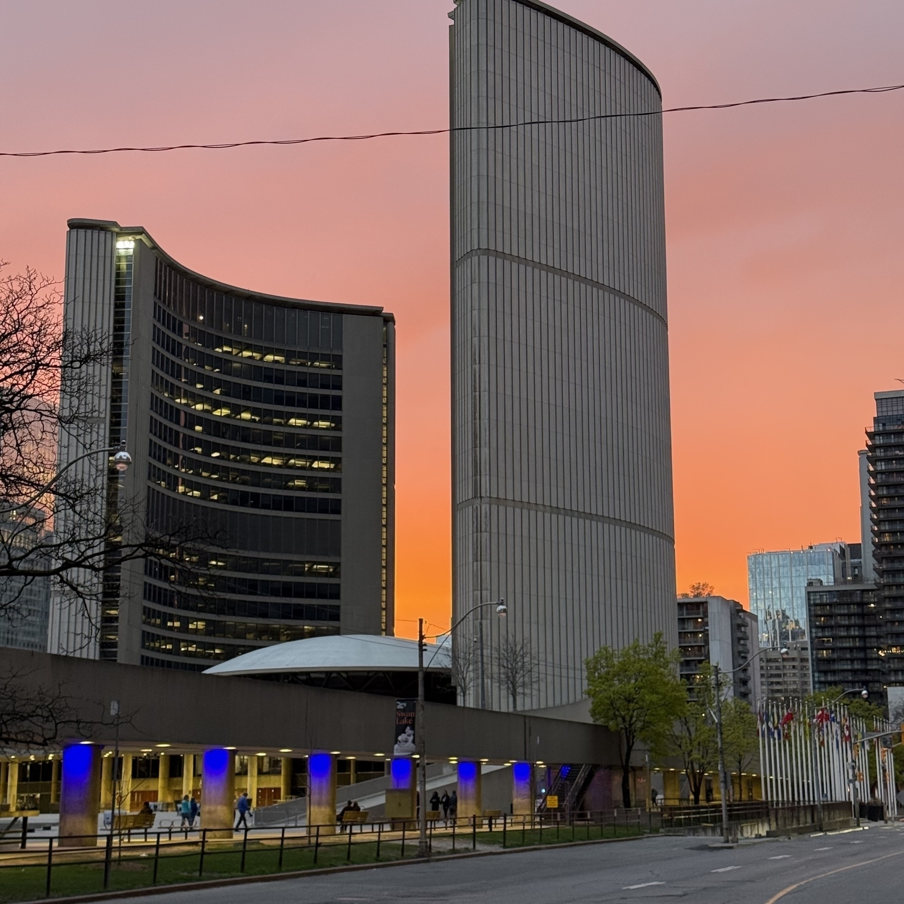 The City of Toronto’s city hall, a tall, modern building with curved facades, stands against a vibrant orange sunset sky, overlooking a plaza illuminated by blue lights.