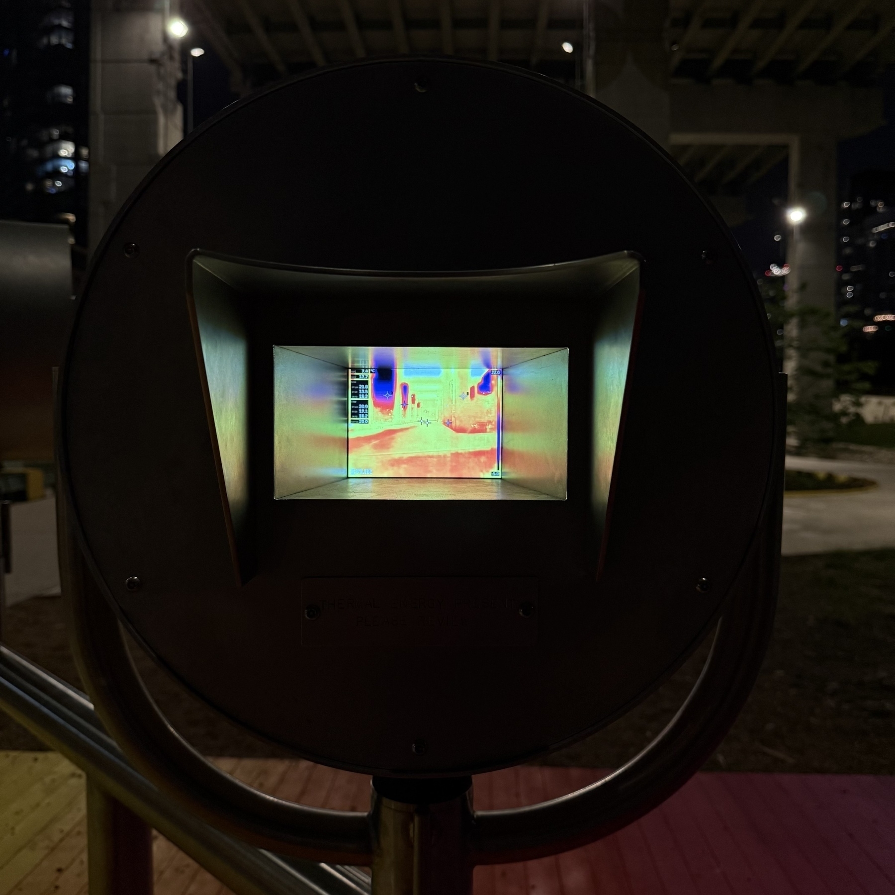 A vibrant, multi-colored light display of a thermal camera is visible through a round, dark viewing apparatus in an urban nighttime setting.