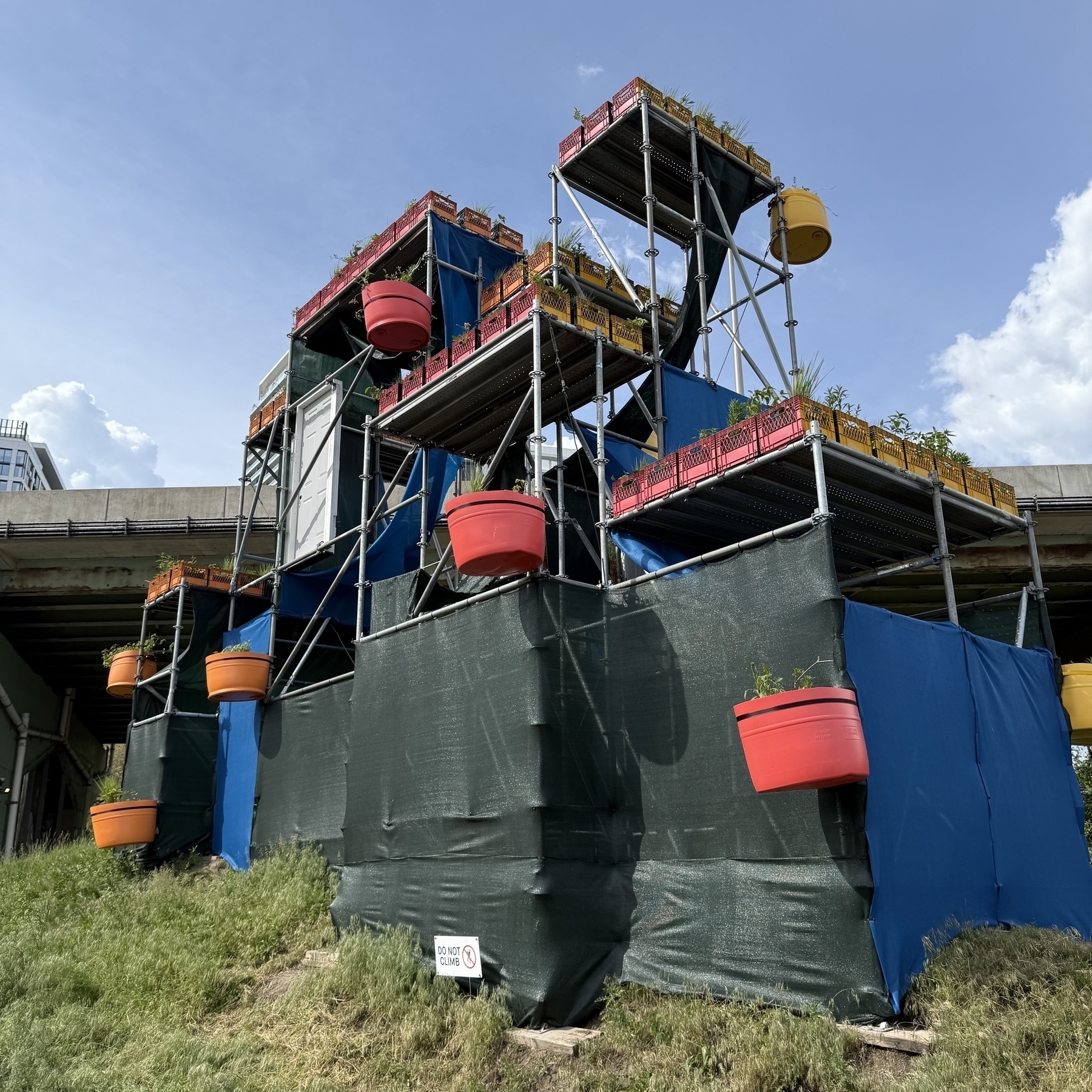 A colorful, multi-level urban garden structure made from large planters and scaffolding is set against a blue sky backdrop.