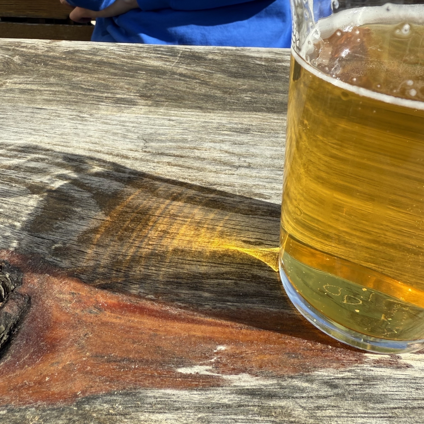 A glass of beer casts a shadow on a wooden table illuminated by sunlight.